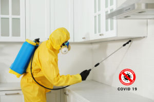 Pest control worker in protective suit spraying insecticide on furniture indoors